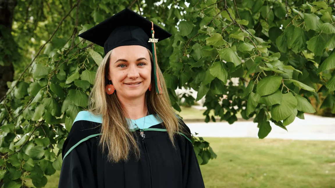 A woman in graduation robes and mortarboard stands in front of green leafy trees, smiling.