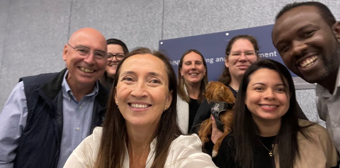 A group of eight people standing indoors pose for a selfie shot.