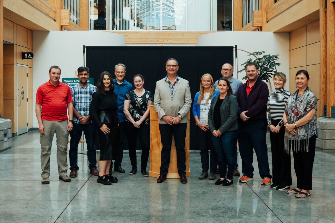 A group of 13 people, including Dr. Indrani Margolin and Dr. Caroline Sanders, stand together in a bright atrium at UNBC. Dr. Bill Owen stands in the center with other faculty members surrounding him.