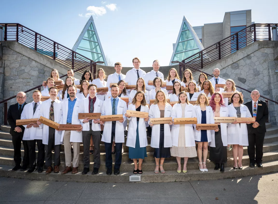 A group of NMP graduates in white coats stand on UNBC's Agora steps.