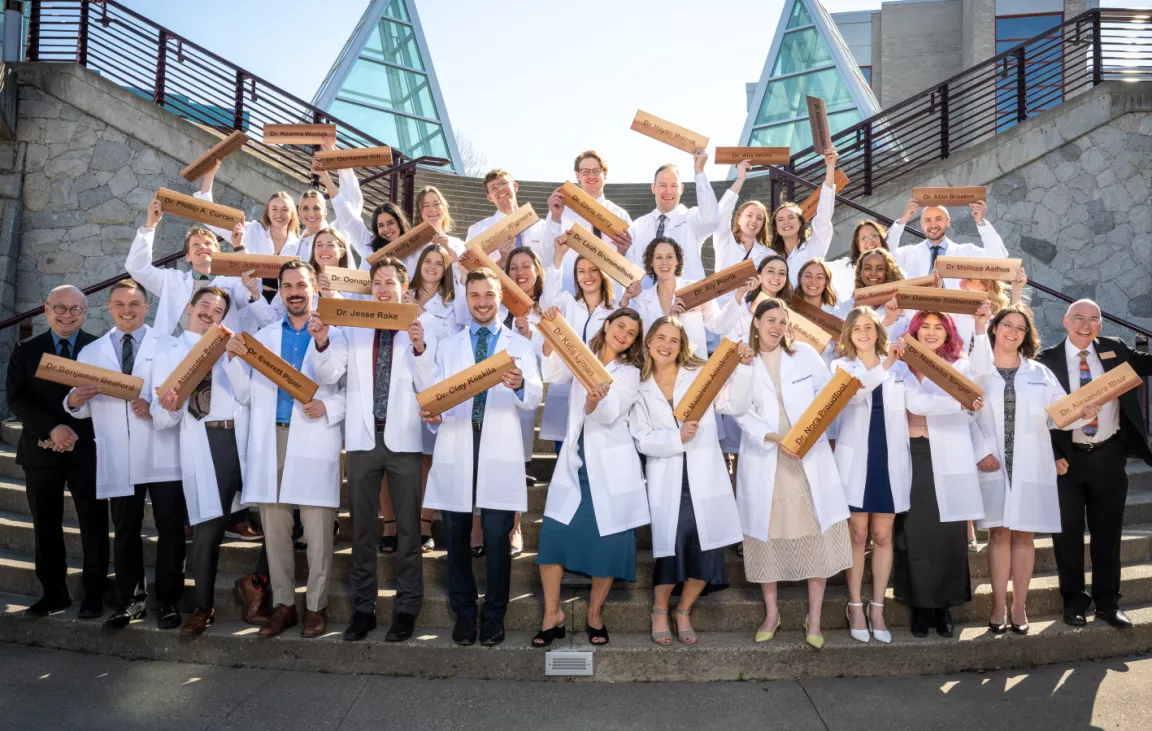 A group of NMP graduates in white coats stand on UNBC's Agora steps.