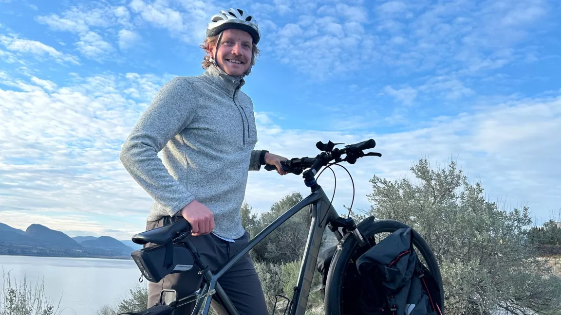 A person in a bike helment stands beside a bike with blue skies and a lake behind them.