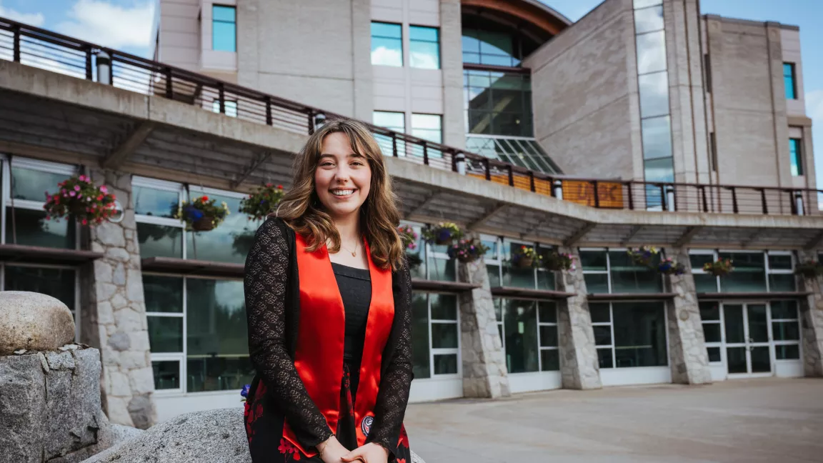 Person wearing black top, black and red skirt, and red satin stole sits on a rock with UNBC Library building in background.