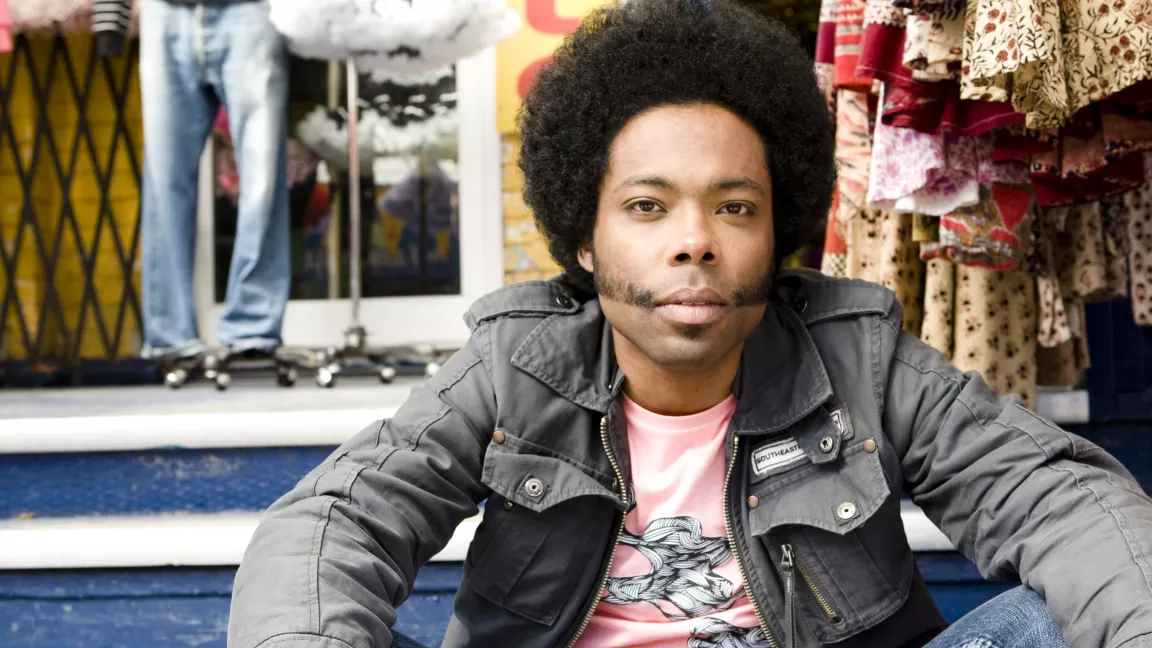 A person with an afro and distinctive sideburns is sitting casually on steps outside a shop. They are wearing a grey jacket over a pink t-shirt, with a calm and relaxed expression. In the background, colorful fabrics and clothing items are displayed on racks.