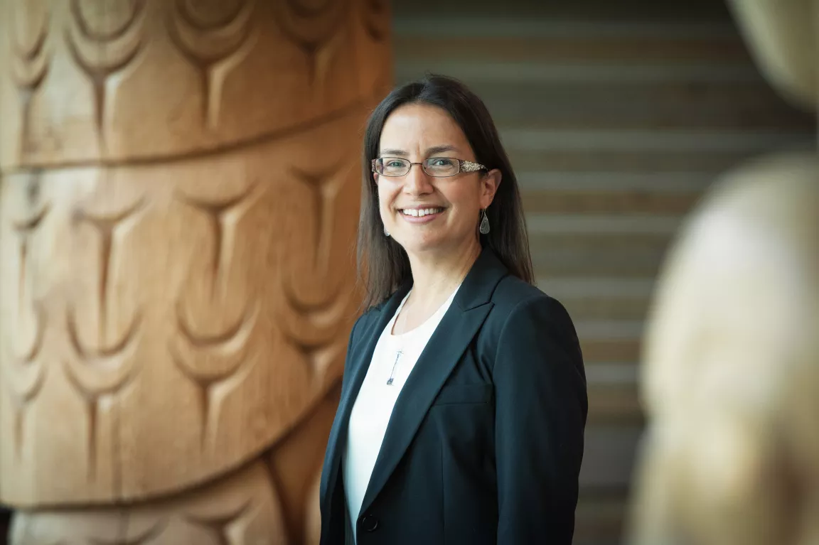 Smiling individual wearing glasses, earrings, white top and dark blazer stands with carved wooden column in background.