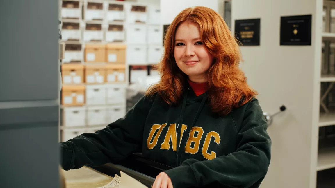 A young woman with red hair wearing a UNBC hoodie smiles while standing in front of archival boxes in the Northern BC Archives.