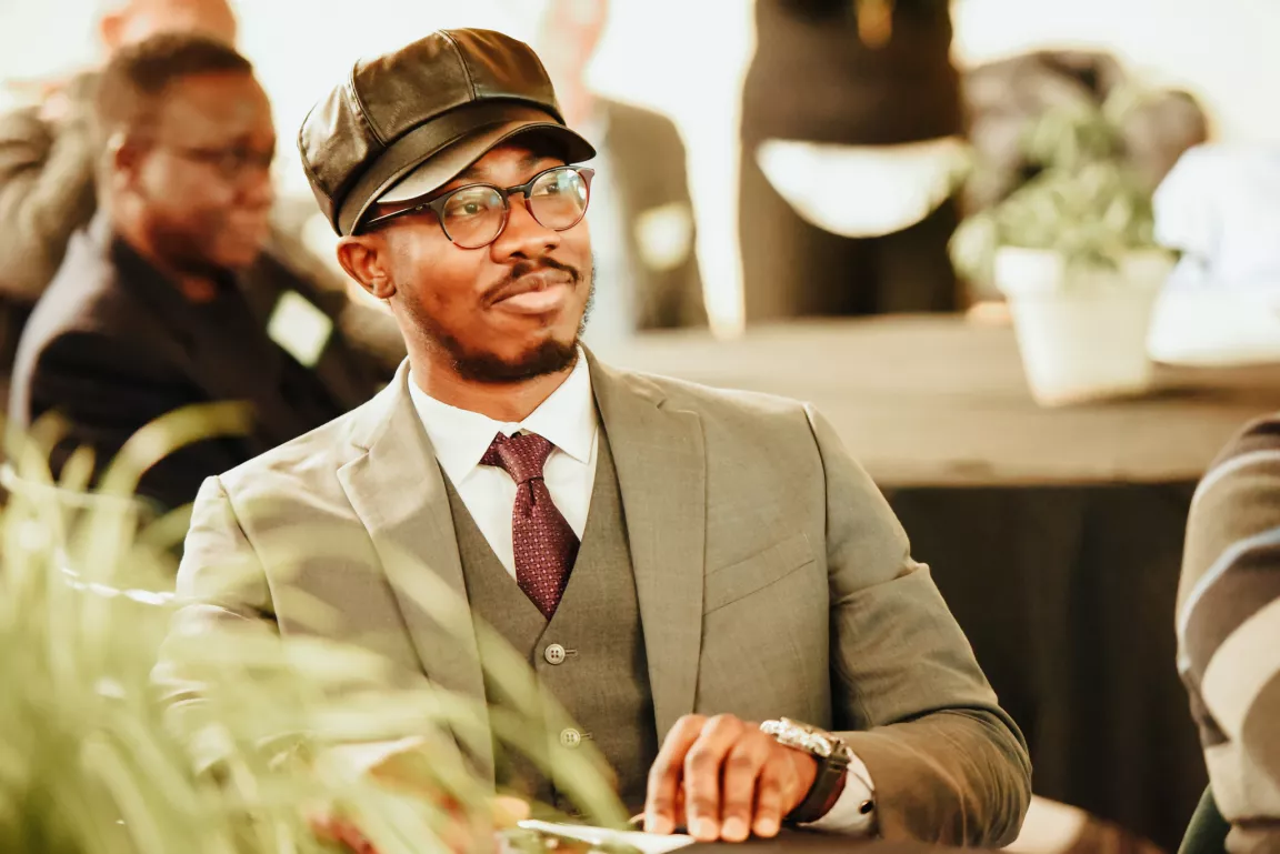 Person wearing black cap, grey suit and burgundy tie sits at a table looking forward