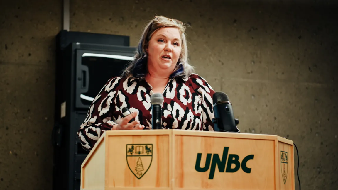 Person wearing patterned blouse stands at a podium with UNBC logo talking to audience.