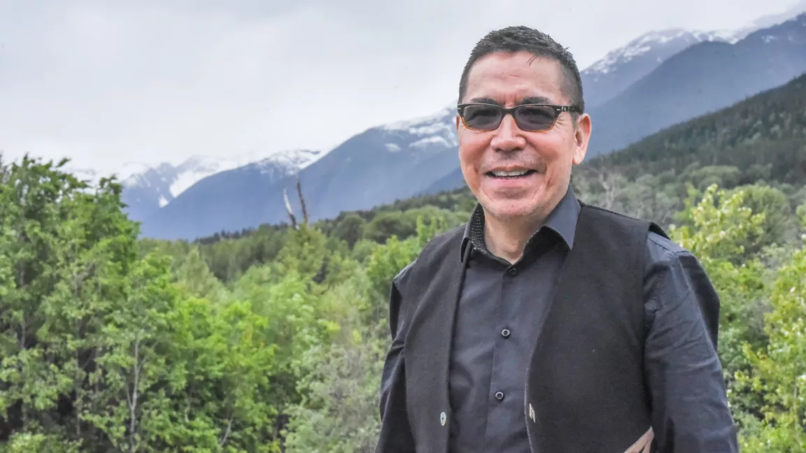 Person wearing dark glass, black shirt and black vest stands in foreground with trees and mountain ridge in background.