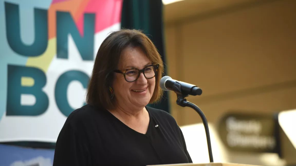 Dr. Margo Greenwood speaks at a podium with a UNBC sign in the background 