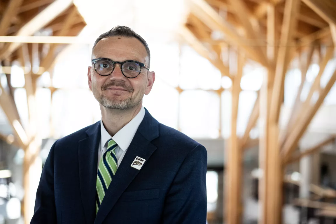 Person wearing glasses, white dress shirt, navy blazer and striped tie stands in an atrium with windows and wood beams in the background.