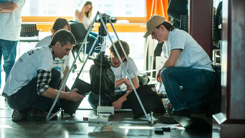 Four people wearing white t-shirts crouch on floor around a small metal structure with a pulley system and weight hanging.