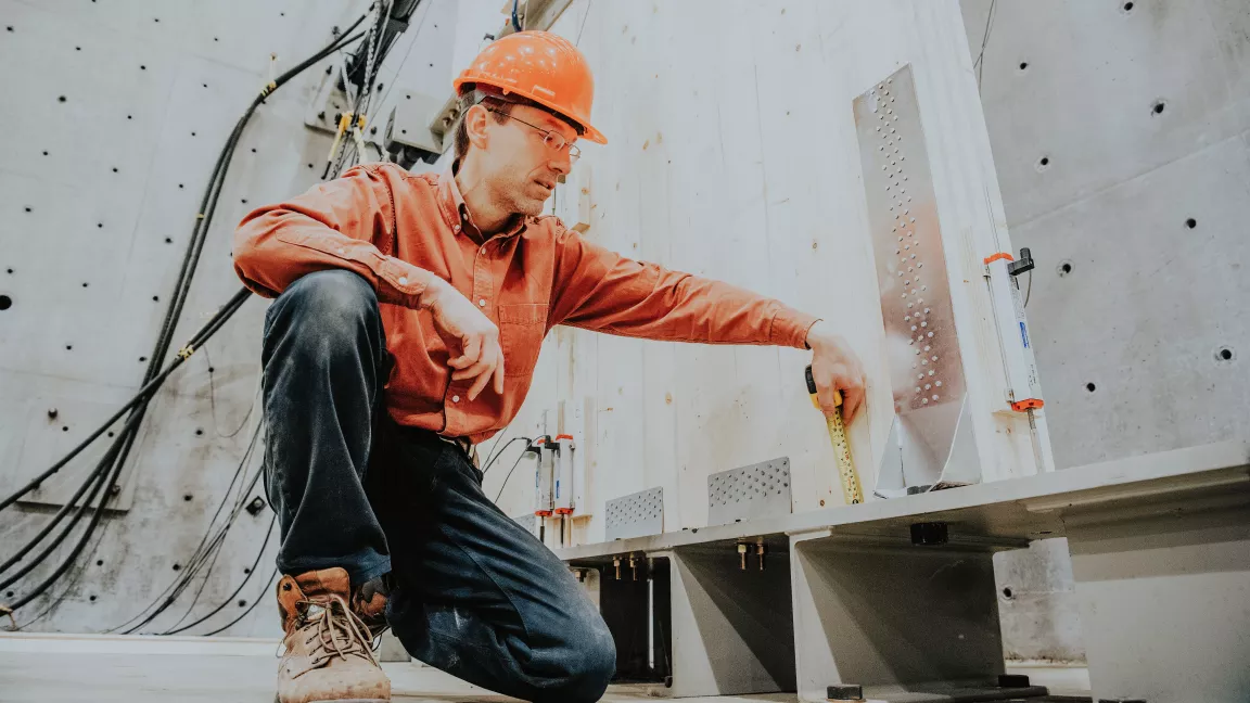 A researcher wearing full personal protective equipment, including a hard had examines a connection next to a strong wall. 