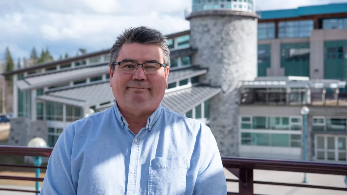 Person wearing blue dress shirt stands outdoors with UNBC's Agora Dining Hall in background.