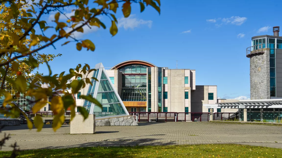 Exterior view of UNBC's Prince George campus showing the Geoffrey R. Weller Library Building with blue sky in background. Tree with fall leaves in foreground.