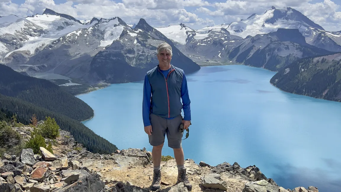 A person wearing a blue and grey jacket and grey shorts stands above a glacial lake with mountains in the background.