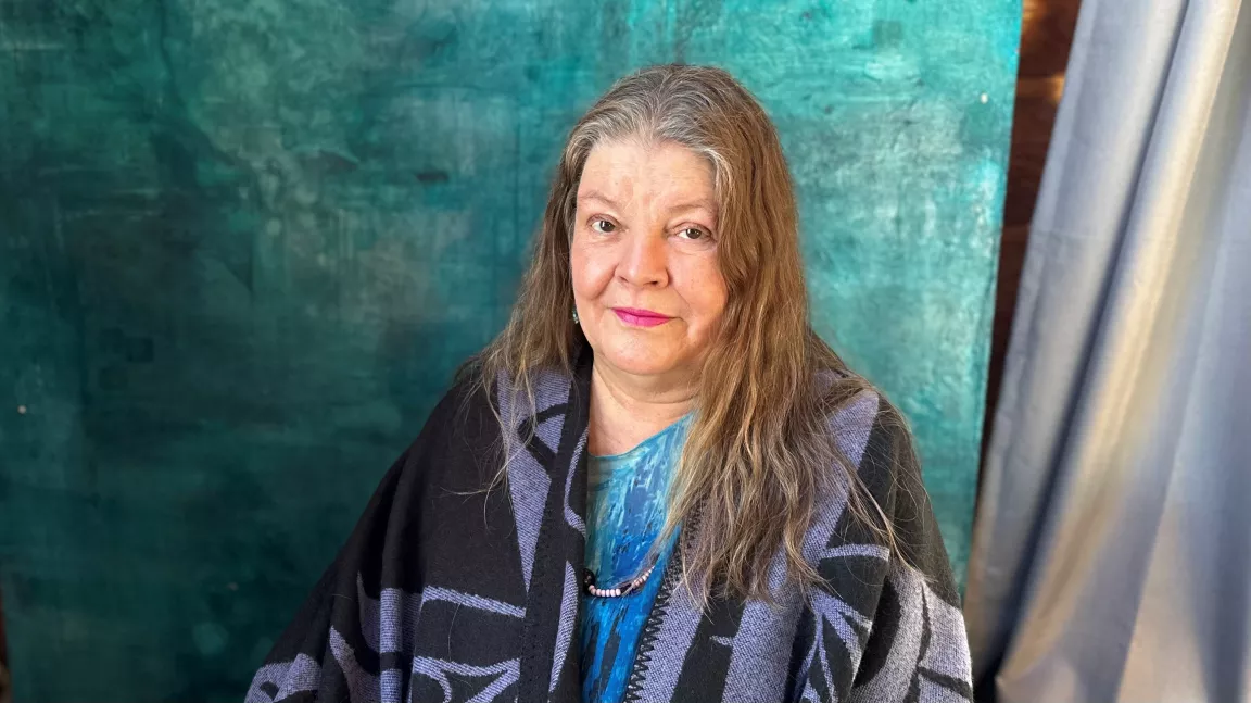 woman wearing blue blouse, and purple wool cover up with black Indigenous art designs on it, sitting in front of green backdrop