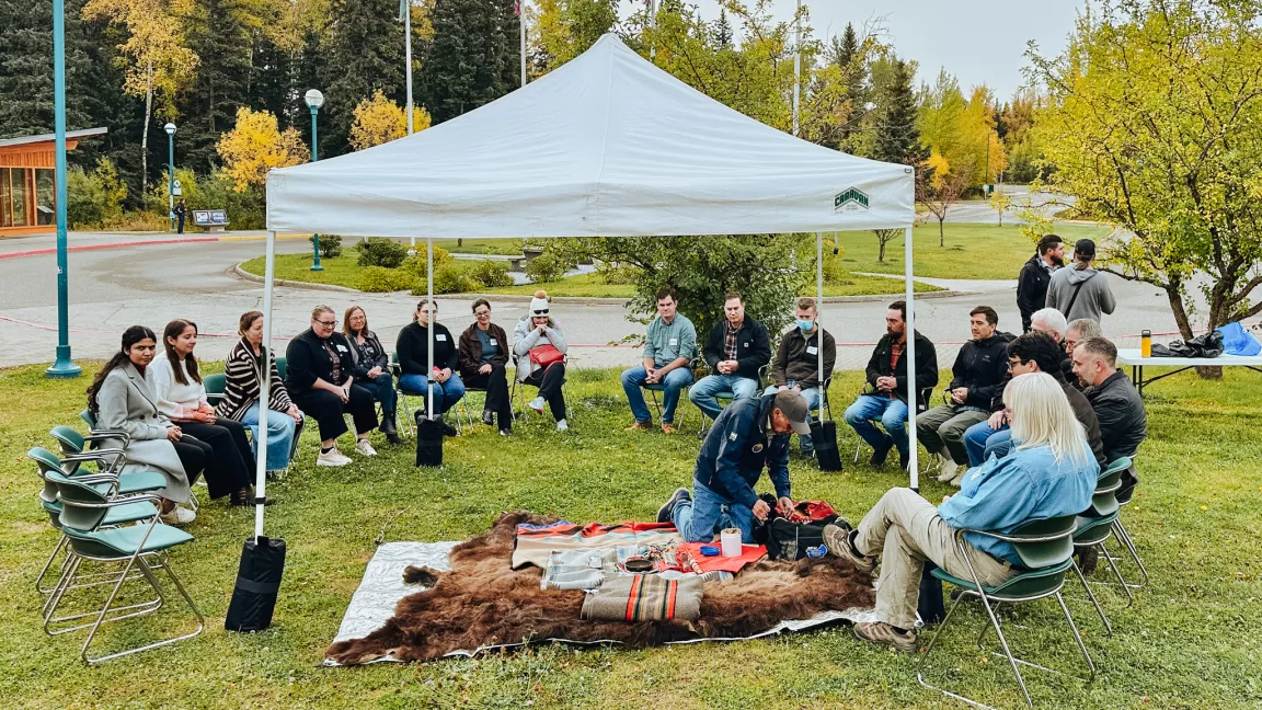 Large group seated on chairs in a large circle outdoors. White canopy in centre and man kneeling on a bear rug reaching into a bag.