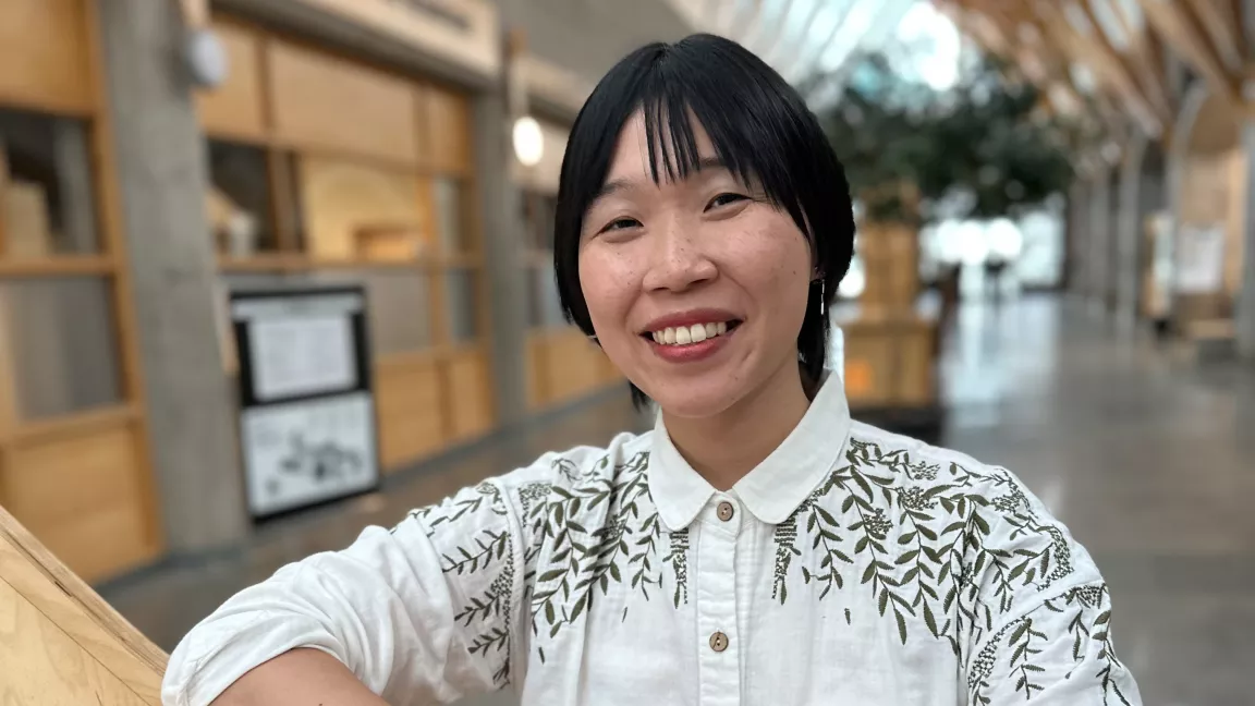 Student wearing white blouse with green embroidery standing in interior hallway.