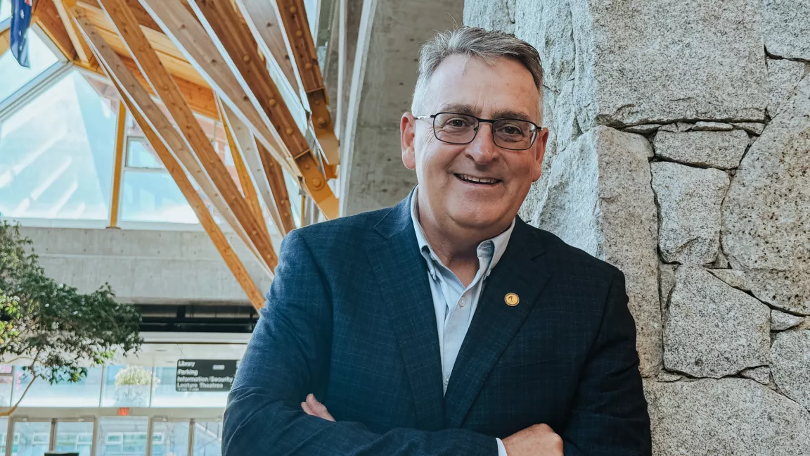 Person wearing blue dress shirt and dark blue blazer with gold pin on lapel leans against a rock wall. Wood beams and windows in background.