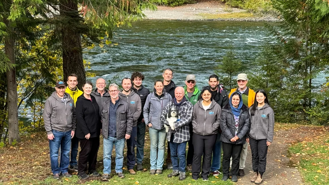 Large group of people standing outdoors with river and fall foliage in background.