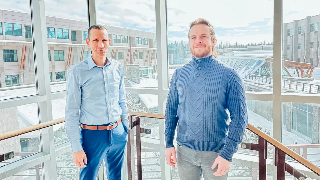 Two people, both wearing blue, stand in a glass building with exterior scenes of UNBC campus in the background.