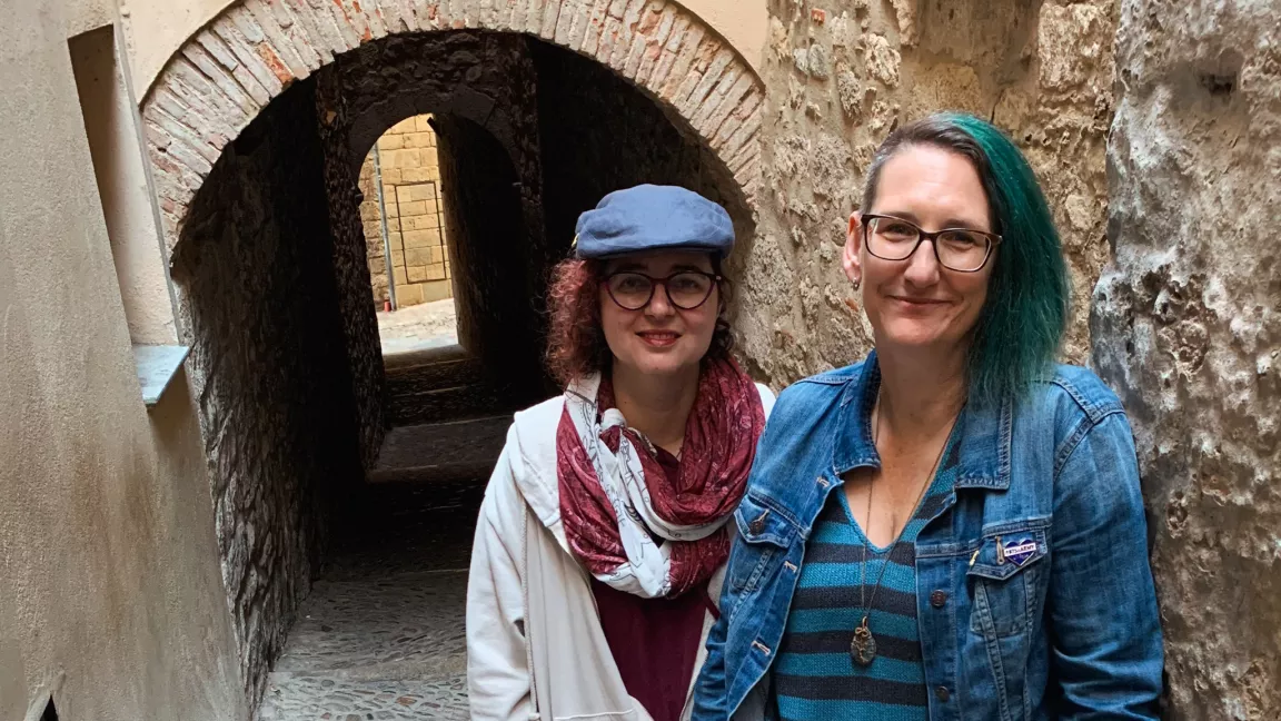 Two people stand in a narrow cobblestone alley with a brick archway in background. Person on right wears blue striped shirt and denim jacket. Person on left wears burgundy top, burgundy and white scarf, white jacket and blue hat.