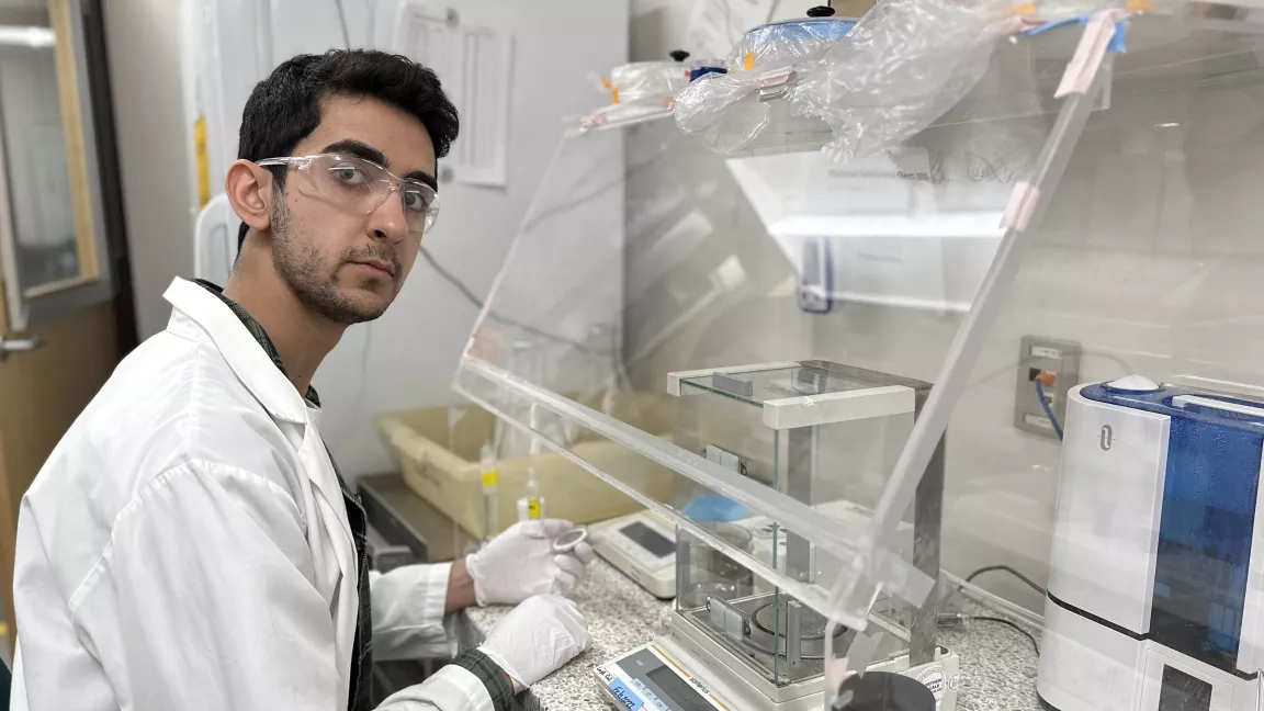 Person in white lab coat with white gloves on sits at a lab table with clear protective hood.