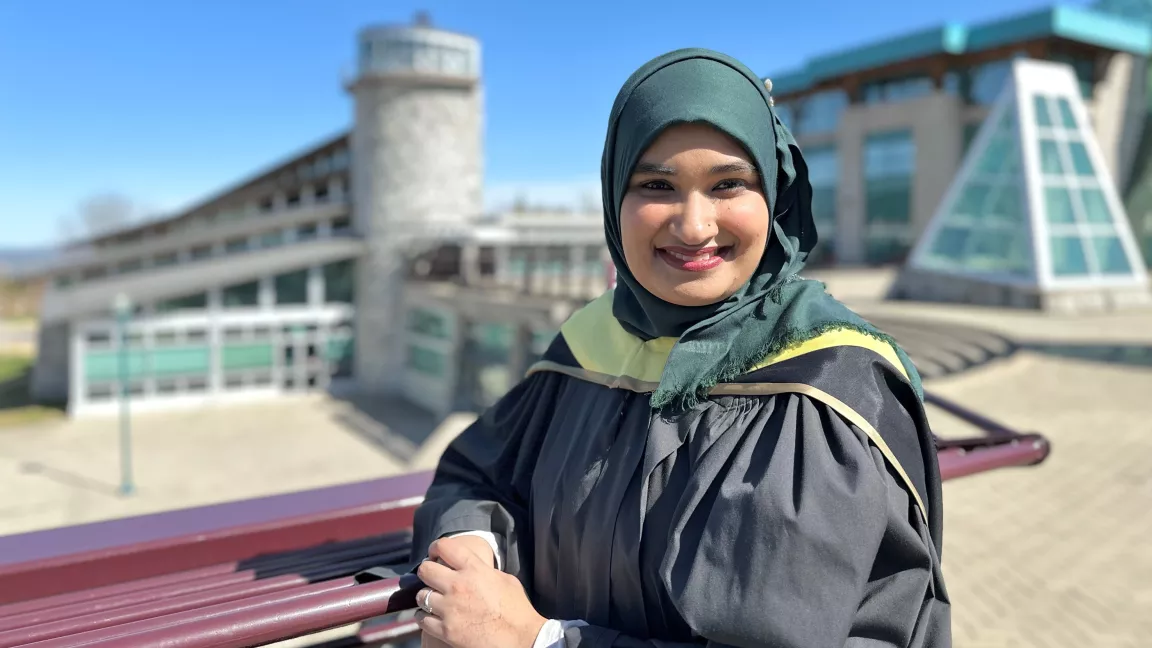 Photo shows exterior shot of person wearing graduation robe and hajib while leaning on burgundy railing.