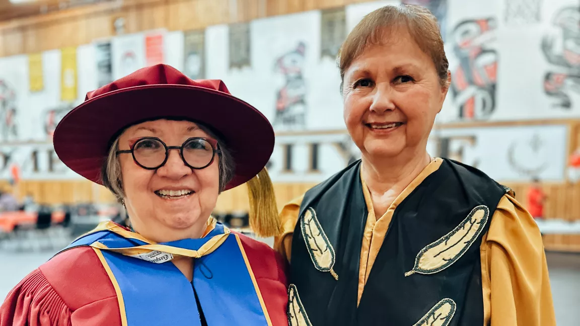 Two people wearing academic regalia smile for the camera. Person on left wears red cap and gown with blue trim. Person on right wears gold robe. Black trim on robe has pattern of gold feathers.