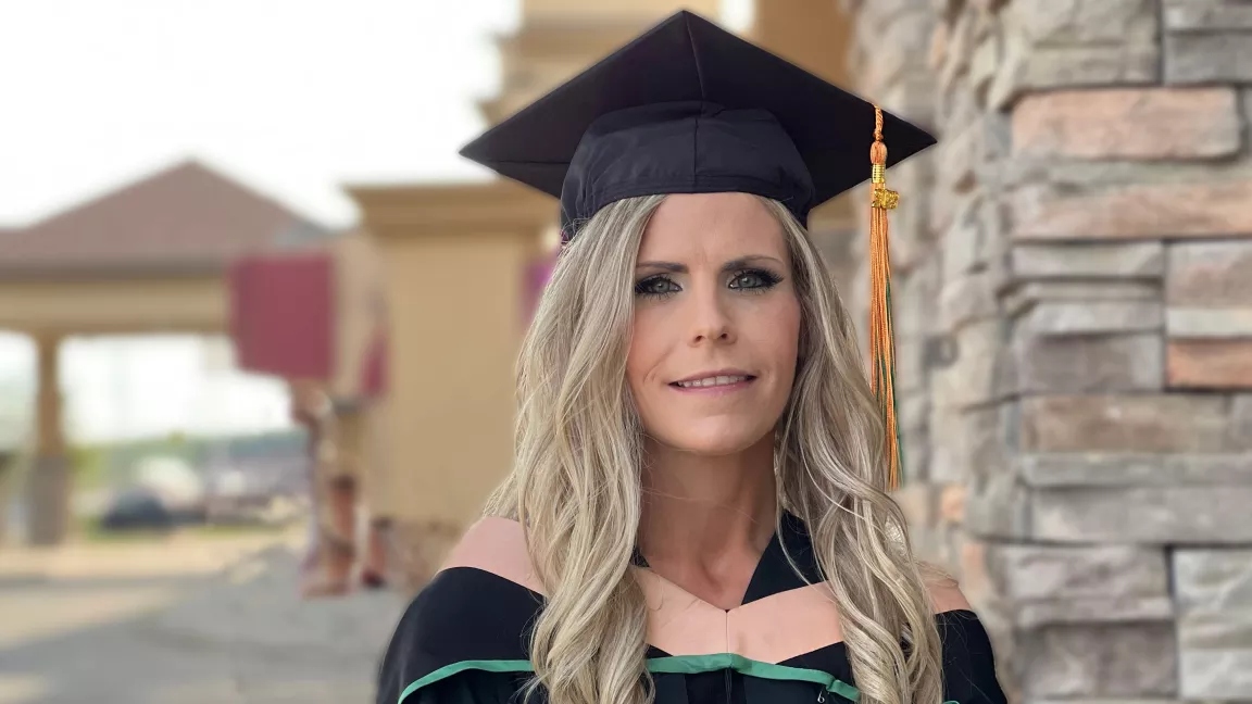 Student wearing academic regalia - black cap and gown with peach and green band - leans against rock column with buildings in background.