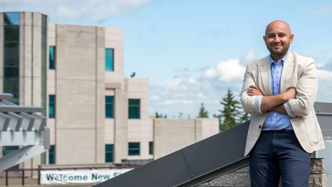 Photo shows student wearing blue dress shirt, light beige blazer and blue slacks leaning against a railing outdoors with building and blue sky in background.