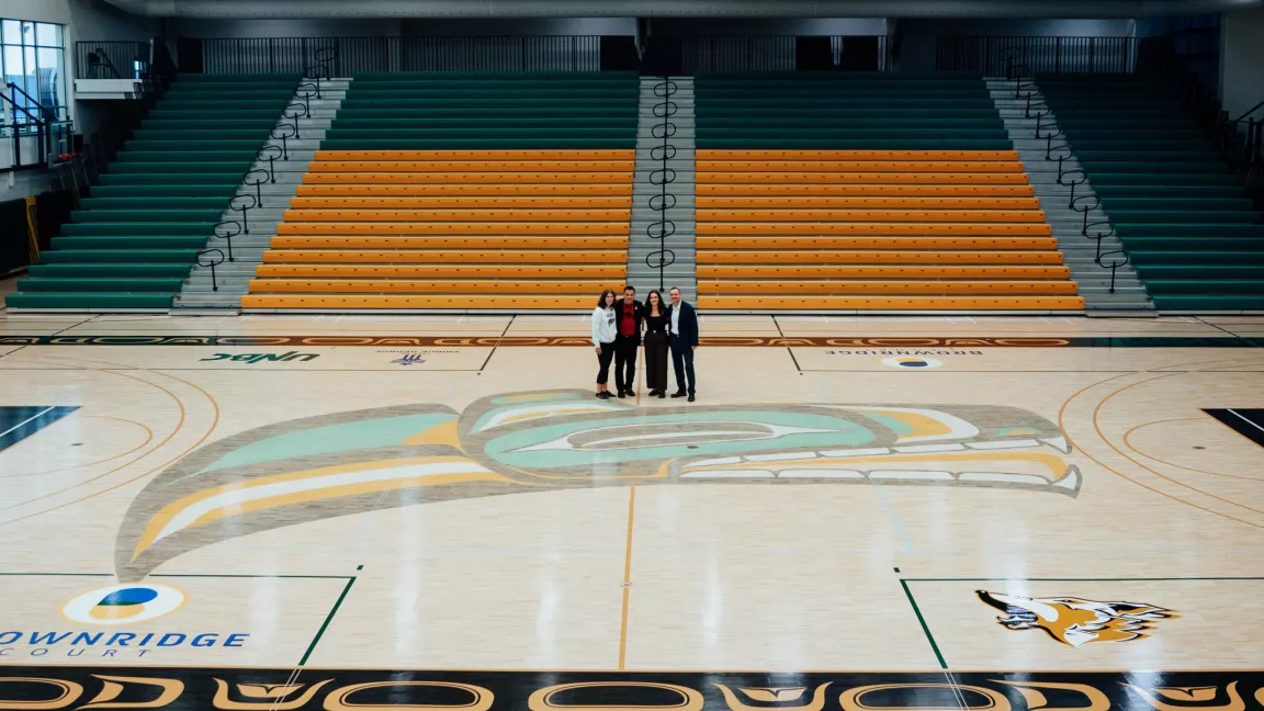 A wide-angle view of the sport floor at UNBC's Brownridge Court, featuring the Timberwolves logo and traditional artwork elements. Four individuals stand at centre court, surrounded by green and gold bleachers.