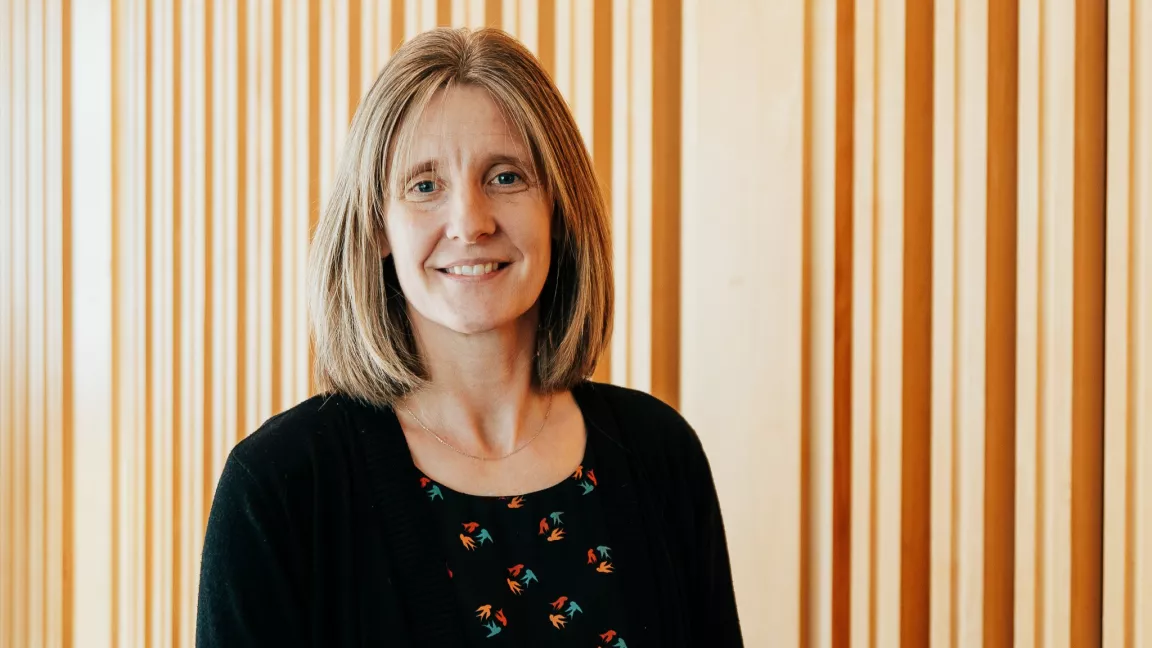 Dr. Sarah Gray, a professor in UBC's Northern Medical Program at UNBC, stands in front of a wooden slatted wall, smiling softly. She has shoulder-length light brown hair and is wearing a black cardigan over a patterned blouse.