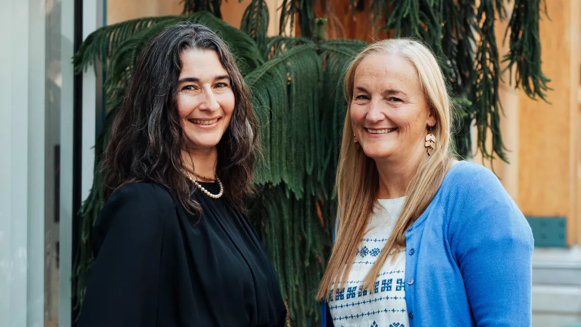 Dr. Indrani Margolin (left) and Dr. Caroline Sanders (right) stand together smiling outdoors in front of greenery. Dr. Margolin is wearing a black top with a pearl necklace, and Dr. Sanders is wearing a blue cardigan and patterned shirt