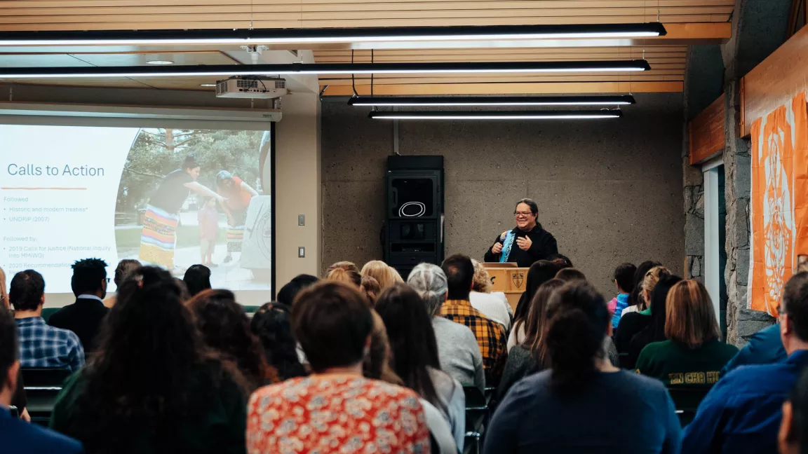 A speaker stands at a podium addressing a seated audience in a room with a presentation screen displaying the title ‘Calls to Action’ and an image of two people interacting outdoors. The audience faces the screen, listening attentively, with orange fabric visible on the wall to the right.