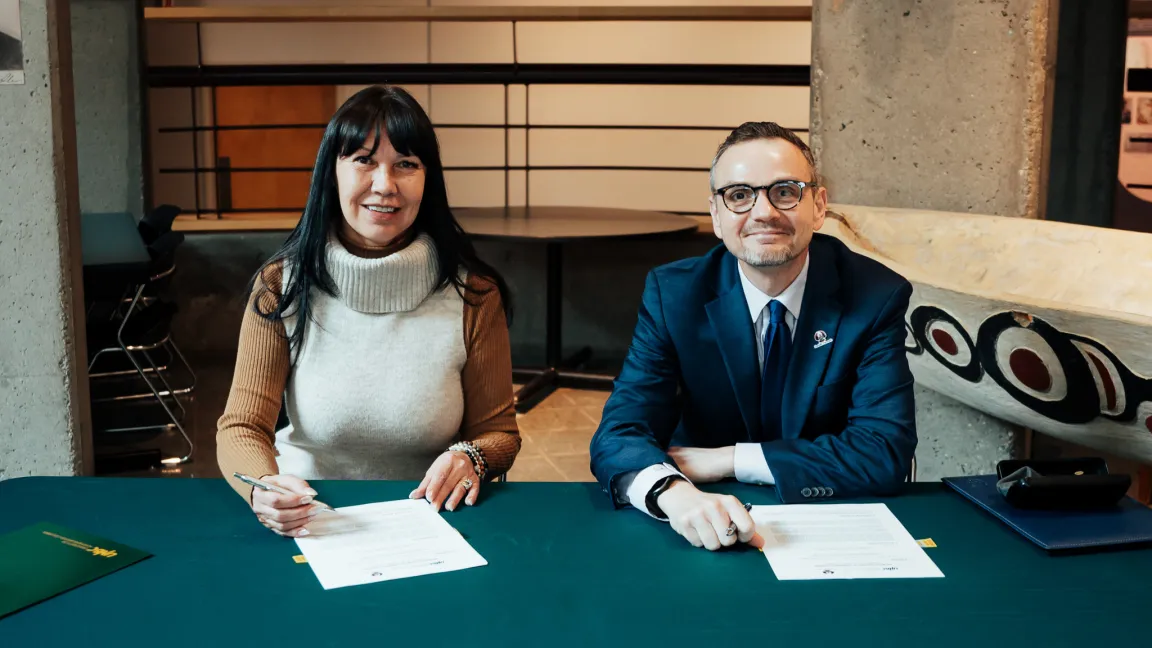 Two people sitting at a table, signing documents. Person on left wears white sweater with cowl neck. Person on right wears blue suit and tie.