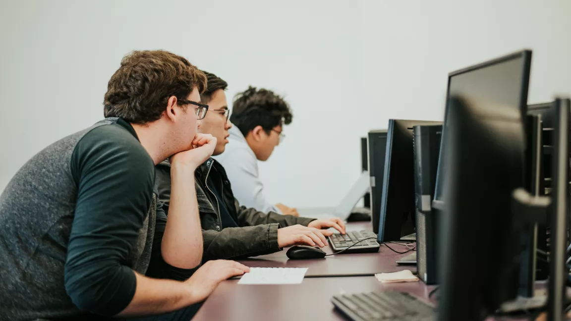 Three students sit in a row at a bank of computer tables. People on left, computer screens and keyboards on right.