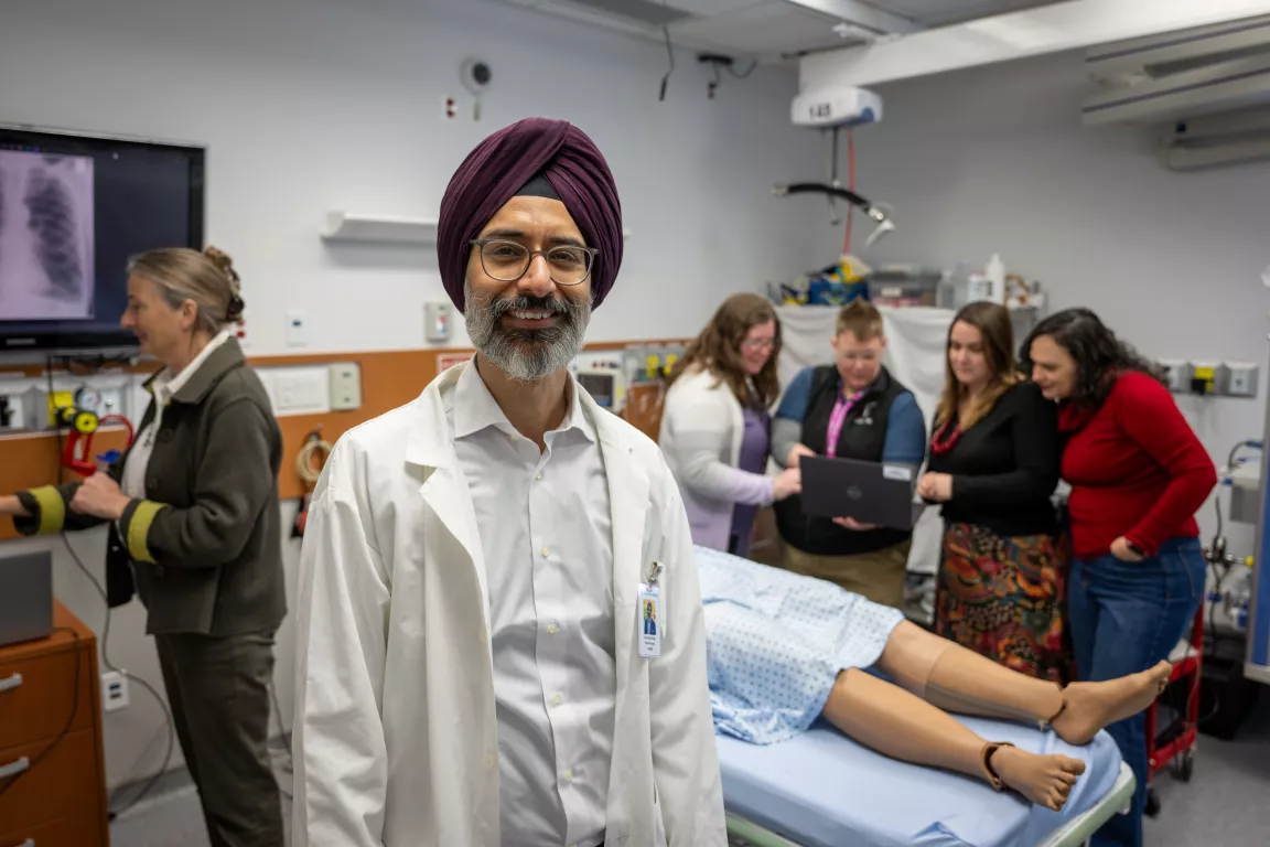 "A man wearing a white lab coat and a maroon turban stands in the foreground of a medical simulation lab, smiling at the camera. Behind him, a group of people is gathered around a hospital bed with a medical mannequin, discussing something on a laptop. A woman in the background is interacting with a screen displaying a chest X-ray."