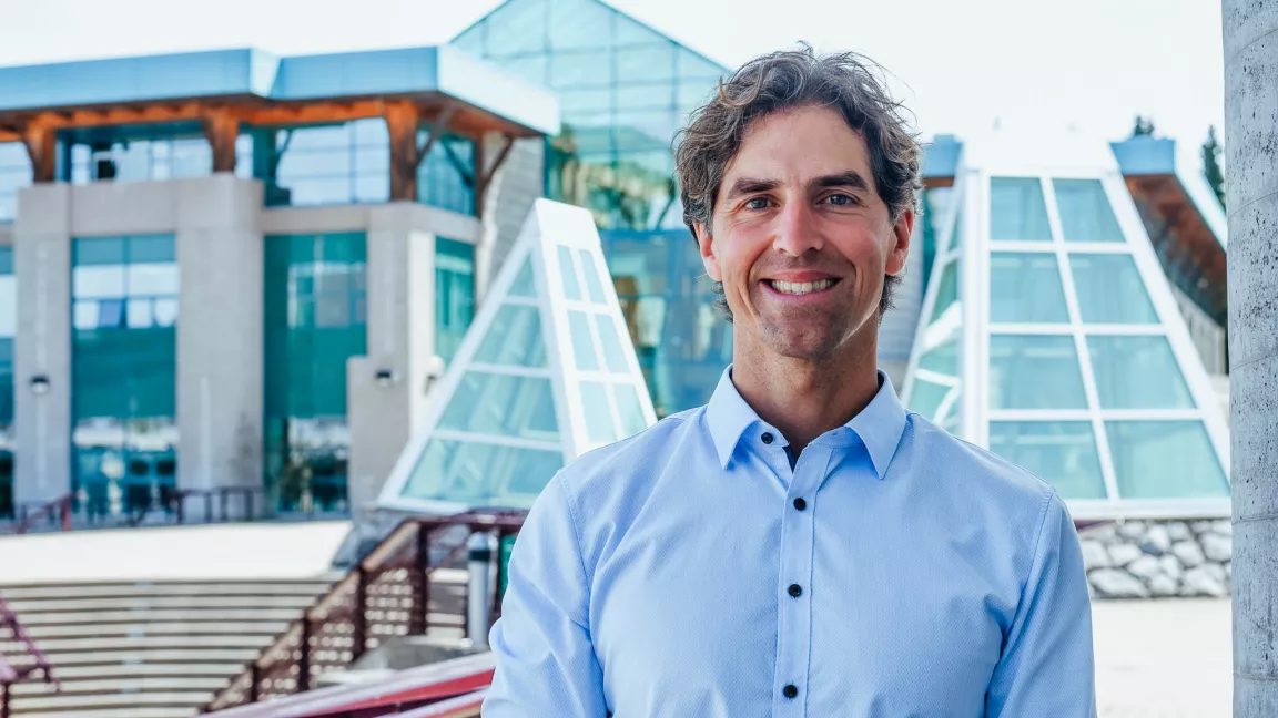 "Dr. Tristan Pearce, Associate Professor in the Department of Geography, Earth, and Environmental Sciences at UNBC, smiling outdoors with a modern glass building in the background.