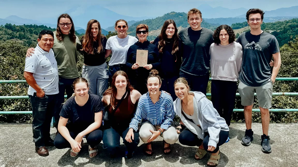 A group of twelve people, including students and faculty from the Guatemala field school, standing and crouching outdoors with a scenic backdrop of mountains and greenery. The person in the centre holds a book, and the group smiles warmly at the camera.