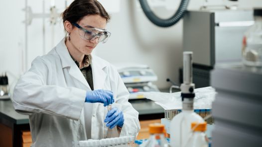 Student wearing white lab coat, safety goggles and blue gloves is working to remove lid on glass test tube.