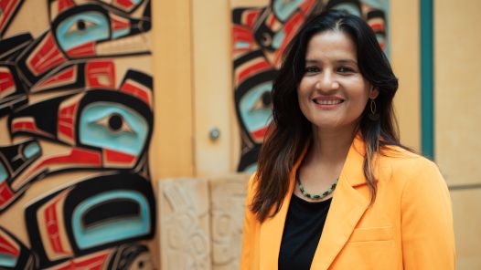 Person wearing black top and orange blazer stands in front of wooden doors featuring Indigenous artwork.