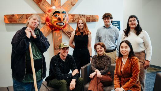 Group of seven people posing indoors with Indigenous artwork in background.