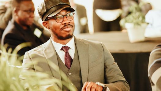 Person wearing black cap, grey suit and burgundy tie sits at a table looking forward