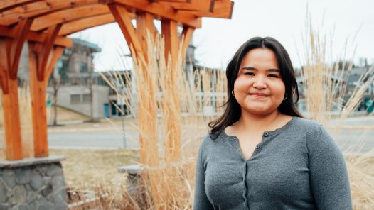 Student wearing a grey sweater stands in front of a wooden archway. UNBC campus in background.