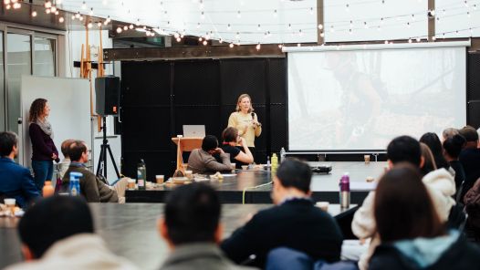 A speaker addresses an audience at UNBC's Wildfire Friday event. The presentation screen shows an image related to wildfire work, and attendees are seated at tables listening attentively. String lights hang above, adding warmth to the indoor setting.