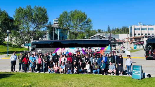 Large group standing outside on grass in front of a bus with UNBC Agora courtyard in background