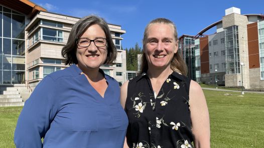 Andrea Gingerich, at left, and Robin Roots stand together on the green lawn with university buildings in the background.