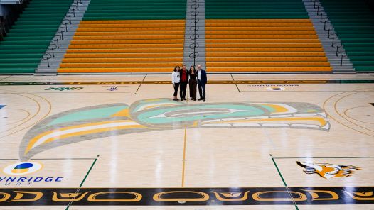 Four people stand at centre court in the Charles Jago Northern Sport Centre gymnasium with Trevor Angus Wolf logo painted on floor. Green and gold seating in background.